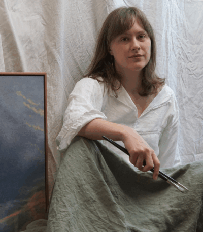 Artist Kaysha Siemens holds paintbrushes while seated beside an Artefex Tempanel-mounted cloud painting in her studio