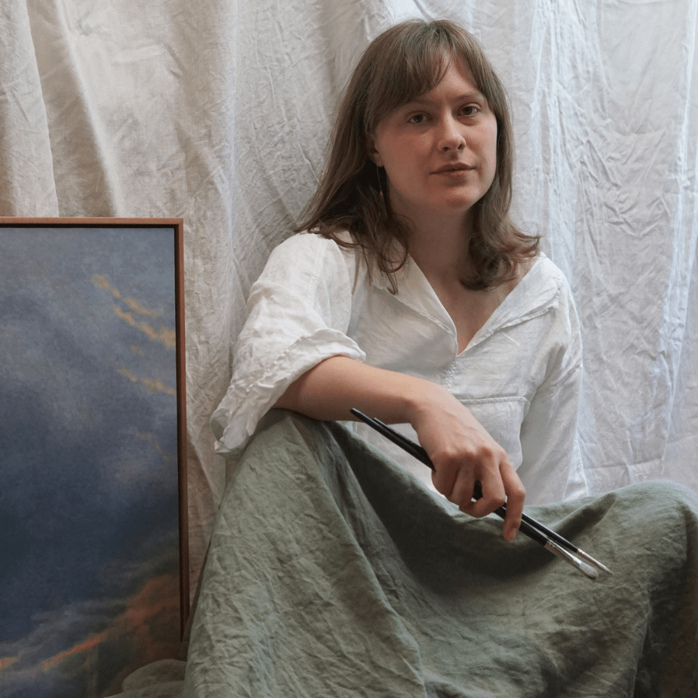 kaysha-siemens-artefex-tempanel-studio Artist Kaysha Siemens holds paintbrushes while seated beside an Artefex Tempanel-mounted cloud painting in her studio
